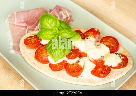 Fladenbrot mit Kirschtomaten, Mozzarella, Basilikumblättern und frischem Schinken auf einem rechteckigen Teller Stockfoto