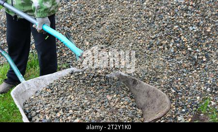 Ein Mann lädt zerquetschten Stein von einem großen Haufen auf dem lokalen Gebiet mit einer Schaufel in eine Gartenkarre und lädt Kies von Hand in eine Schubkarre Stockfoto