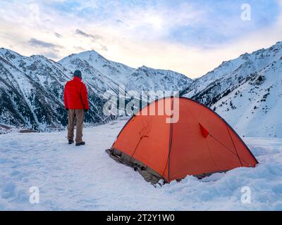 Ein männlicher Tourist steht früh am Morgen in den schneebedeckten Bergen nahe dem roten Zelt, in dem er die Nacht verbrachte. Ein Reisender in einer roten Daunenjacke trave Stockfoto