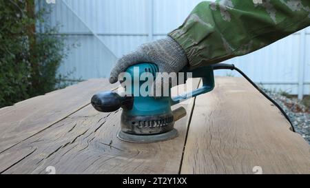 Zimmermann in Handschuhen arbeitet mit einem Schleifgerät in einem grünen Gehäuse mit rundem Schmirgelaufsatz auf Polieren massiver Eichenbretter in einer Werkstatt im Freien Stockfoto