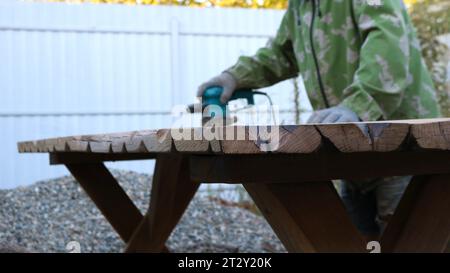 Handgefertigter Massivholztisch mit strukturierten Tischplattenkanten im Oberflächenveredelungsprozess in der Werkstatt im Freien mit Zimmermann in Unschärfe Stockfoto