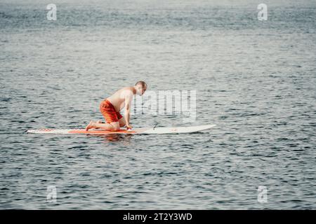 Aktiver reifer männlicher Paddler mit seinem Paddleboard und paddeln im Sommer auf dem Meer. Ein glücklicher Seniorenmann steht mit einem SUP-Brett. Stand-Up-Paddle-Boarding - Stockfoto