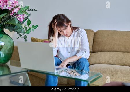 Müde Frau mittleren Alters, die an Kopfschmerzen leidet, sitzt auf Sofa mit Laptop Stockfoto