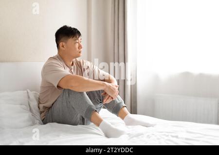Einsamer asiatischer Mann mittleren Alters, der Pyjamas trägt und auf dem Bett sitzt Stockfoto