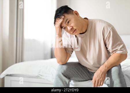 Depressiver chinesischer Mann mittleren Alters, der Pyjamas trägt und auf dem Bett sitzt Stockfoto