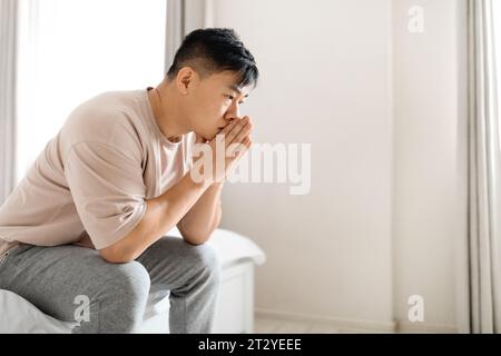 Nachdenklicher, nachdenklicher, reifer asiatischer Mann, der Pyjamas trägt und auf dem Bett sitzt Stockfoto