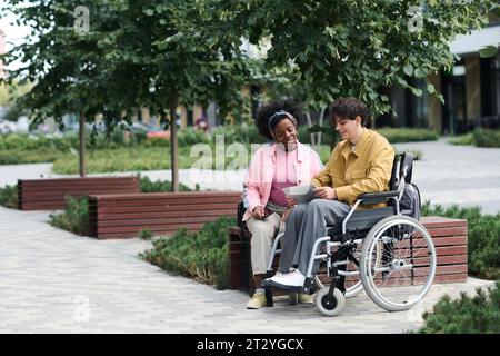 Mann mit Behinderung verbringt Zeit im Freien mit einem Freund, sie benutzen ein digitales Tablet und schauen sich lustige Videos an Stockfoto