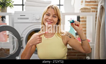 Junge blonde Frau, die Kleidung wäscht und auf Waschmittelbeutel im Waschraum zeigt Stockfoto
