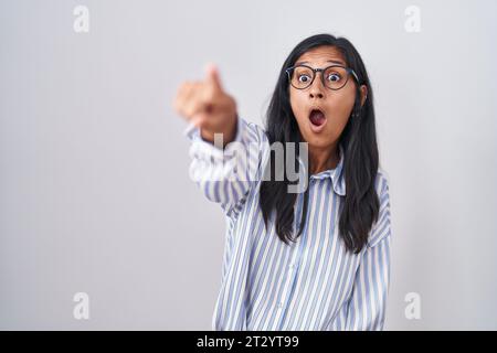 Die junge hispanische Frau trägt eine Brille, die mit dem Finger nach vorne zeigt, der offene Mund verblüfft, etwas auf der Vorderseite Stockfoto