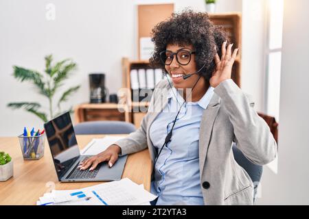 Schwarze Frau mit lockigem Haar, die im Büro ein Callcenter-Agenten-Headset trägt, lächelt mit Hand über Ohr und hört Gerüchte oder Klatsch zu. Taub Stockfoto