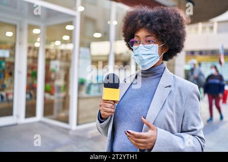 afroamerikanische Reporterin trägt eine medizinische Maske und benutzt ein Mikrofon, das auf der Straße arbeitet Stockfoto