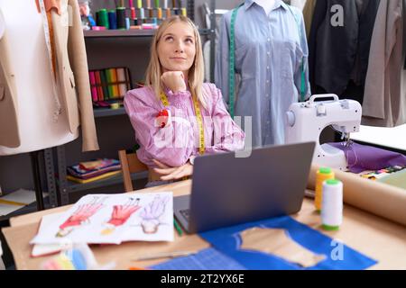 Junge weiße Schneiderin Designerin, die Laptop benutzt, konzentrierte sich auf Zweifel mit Finger am Kinn und schaute sich wundernd auf Stockfoto