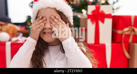 Entzückendes hispanisches Mädchen, das die Augen mit Händen bedeckt, die zu Hause am weihnachtsbaum auf dem Boden sitzen Stockfoto