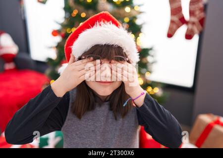 Entzückendes hispanisches Mädchen, das die Augen mit Händen bedeckt, die zu Hause am weihnachtsbaum stehen Stockfoto