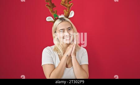 Junge schöne hispanische Frau lächelt selbstbewusst mit Rentier-weihnachtshut über isoliertem roten Hintergrund Stockfoto