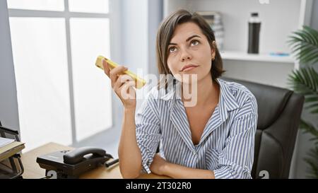 Ernsthafte und doch entspannte junge, schöne hispanische Geschäftsfrau, die in tiefgründige Gedanken vertieft ist, sich auf ihr Smartphone an ihrem Schreibtisch konzentriert und verkörpert Stockfoto