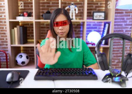 chinesische Frau mittleren Alters, die eine Virtual-Reality-Brille trägt, hört auf zu singen mit der Handfläche. Warnausdruck mit negativer und ernster Geste o Stockfoto