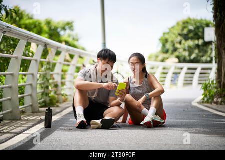 Junge asiatische Paare, die sich Handyfotos anschauen, während sie eine Pause während des Outdoor-Trainings machen Stockfoto