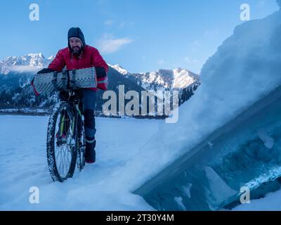 Im Winter fährt ein Mann mit dem Fahrrad in den Bergen. Radtour im Winter. Schlittschuhlaufen auf dem Eis eines gefrorenen Bergsees. Früher Wintermorgen hoch im Stockfoto