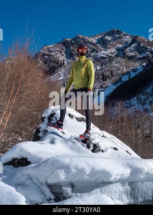 Ein bärtiger Sportler steht im Winter auf einem Felsen hoch in den Bergen. Ein Typ mit Sonnenbrille und leichten Klamotten trainiert im Winter draußen. Stockfoto