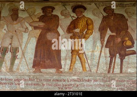 Detail aus dem 16. Jahrhundert Danza Macabra (Tanz des Todes) Freskenfries: Lächelnde Skelette mit doppelt amputiertem Bettler (rechts), Soldat in Rüstung (links), Dandy oder eitler junger Mann (rechts in der Mitte) und Mann in flacher Mütze, jeder Sterbliche durchbohrt von Pfeil des Todes; der Dandy trägt ein zerschnittenes Wimperntuch, einen Kofferschlauch, Puffärmel, einen Spitzenkragen und eine Feder im Hut. Gemalt von der italienischen Künstlerin Simone II. Baschenis (ca. 1495–1555) an der Außenwand der Chiesa di San Vigil in Pinzolo, Trentino-Südtirol, Italien. Stockfoto
