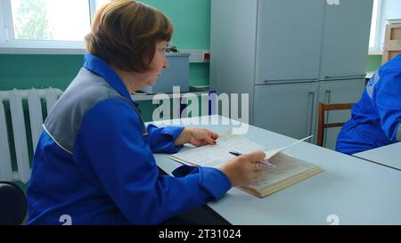 Ältere Frau macht Notizen im Labor. Clip. Weiblicher Dispatcher in Uniform schreibt in Log. Dispatcher zeichnet Daten auf Geräten im technischen Protokoll auf Stockfoto