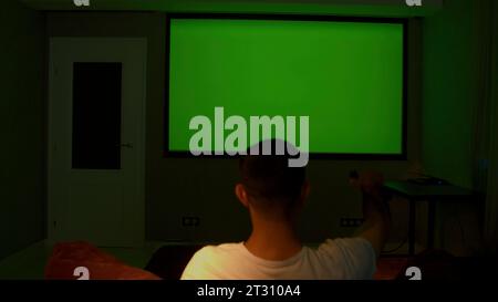 Ein Mann, der auf der Couch sitzt. Medien. Ein erwachsener Mann, der in einem Zimmer ruht und auf einen grünen Bildschirm blickt. Stockfoto