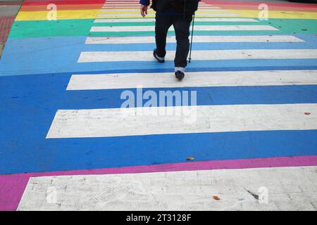 Ein Mann mit einem Stock überquert eine Straße. Die Zebrakreuzung ist in Regenbogenfarben gemalt Stockfoto