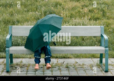 Ein kleines Mädchen versteckt sich vor dem Regen unter einem Regenschirm. Sie sitzt auf einer Bank. Das Mädchen trägt pinkfarbene Gummistiefel und Regenhosen. Es gibt ein g Stockfoto