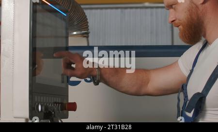 Man klickt auf das Bedienfeld. Kreativ. Mann mit Bart in Arbeitsuniform in der Fabrik. Man stellt das Bedienfeld von Industriemaschinen ein Stockfoto