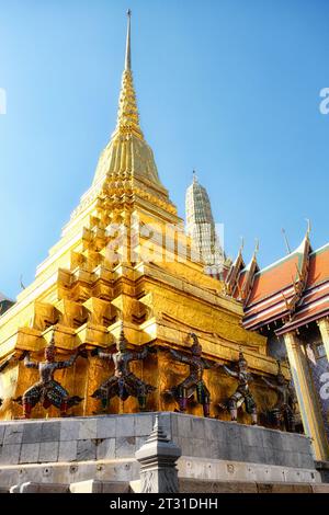 Ein goldener Tempelturm in Thailand, unterstützt von Statuen von mythischen Kreaturen, die mit bunten Mosaikfliesen verziert sind, klarem blauem Himmel. Stockfoto