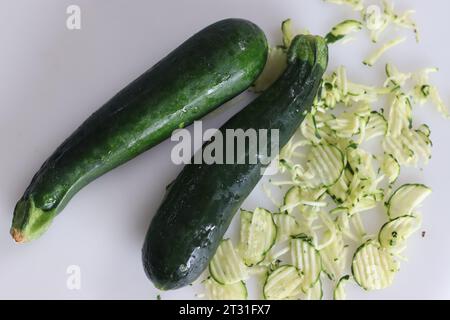 Üppige, grüne Zucchini, eine Zucchini-Spezies, die Gemüse oder auch Babymark genannt wird, fotografiert auf weißem Hintergrund zusammen mit geschredderter Zucchini. Stockfoto