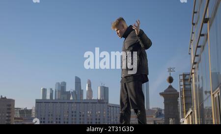Guy tanzt cool auf dem Hintergrund der modernen Stadt. Aktion. Junger Mann tanzt mit Blick auf die Stadt an sonnigen Tagen. Moderner Tanz des jungen Mannes am Glas Stockfoto