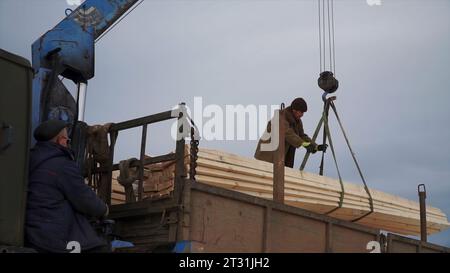 Männer entladen Kran mit Holz. Clip. Arbeiter, die Holz mit einem Kran entladen. Kran mit Holzbalken wird mit Arbeiter in den Lkw abgesenkt Stockfoto