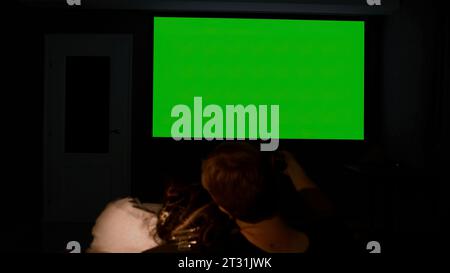 Zu Hause auf einem großen Projektor ansehen. Medien. Ein junges Paar, das sich auf der Couch umgibt und auf einen grünen Bildschirm blickt. Stockfoto