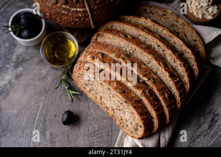 Gebackenes Sauerteigbrot aus Vollkornmehl und Kürbiskernen auf einem Gitter, Olivenöl und schwarze Olive auf einem rustikalen Holztisch. Handwerkliches Brot. Stockfoto