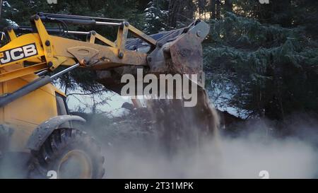 Traktor schaufelt Schnee und Erde im Wald. Clip. Der Traktor räumt im Winter Platz im Wald. Der Traktor gräbt und gießt Erde in verschneiten Wäldern aus Stockfoto