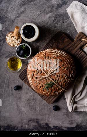 Gebackenes Sauerteigbrot aus Vollkornmehl und Kürbiskernen auf einem Gitter, Olivenöl und schwarze Olive auf einem rustikalen Holztisch. Handwerkliches Brot. Stockfoto
