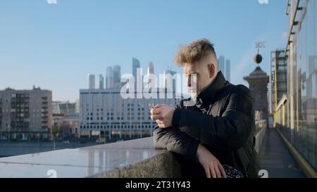 Junger Mann schaut von der Brücke aus auf die Stadt. Aktion. Mann auf Brücke mit Blick auf die sonnige moderne Stadt. Mann in Jacke steht an sonnigem Tag auf der Brücke Stockfoto