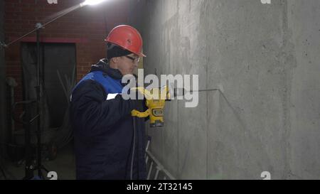 Builder in Uniform bohrt Löcher in der Wand. Clip. Der Servicetechniker macht mit dem Bohrer ein Loch. Bauarbeiter mit Bohrer macht ein Loch in einem neuen Raum Stockfoto