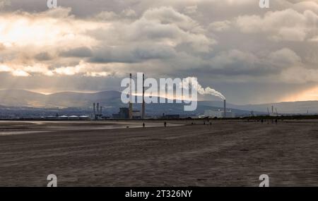 Am Dollymount Strand Beach bricht Licht durch dunkle Wolken. Poolbeg Schornsteinstapel und Kraftwerk in der Ferne. Dublin, Irland Stockfoto