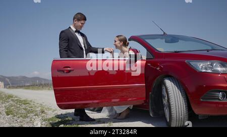 Jungvermählte beim Cabrio, Gefühle der Zärtlichkeit. Aktion. Ein Mann im Anzug hilft der Braut, das rote Auto zu verlassen. Stockfoto