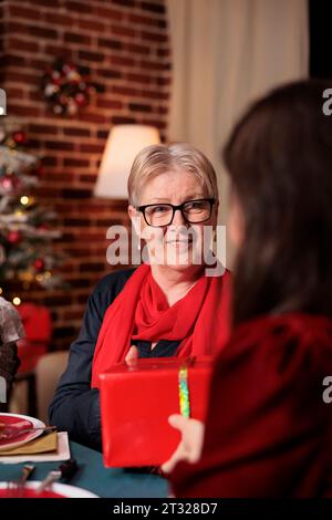 Frau, die ihrer Mutter bei der weihnachtsfeier ein Geschenk schenkt und sich während der dezemberfeier fröhlich fühlt. Die Familie tauscht Geschenke aus und genießt eine schöne Zeit zu Hause. Stockfoto