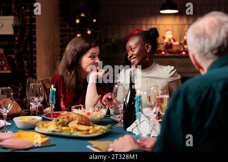 Verschiedene Freunde treffen sich zu weihnachten zum Abendessen und feiern die dezember-Feiertage zu Hause. Menschen, die feiern, umgeben von festlichen Dekorationen und Weihnachtsbaum, glücklich fühlen. Stockfoto