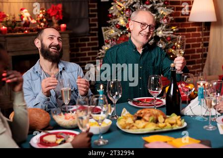 Ein Senior Mann, der am weihnachtsabend mit einem Glas Wein Toast trinkt, um die dezember-Feiertage mit Freunden und Familie zu feiern. Großvater hält eine Rede am Tisch, spritziger Alkohol. Stockfoto