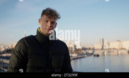 Mann mit kurzen lockigen Haaren, der durch die Stadt läuft. Aktion. Konzept des Reisens, Mann in wark Jacke, die mit dem Fluss im Hintergrund läuft. Stockfoto