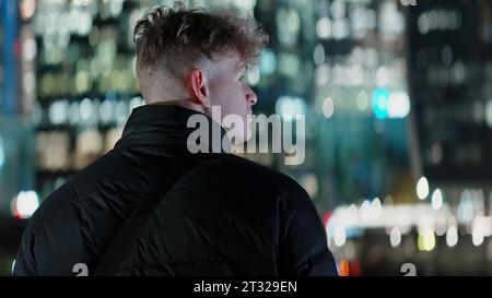 Mann mit kurzen lockigen Haaren, der nachts in die Stadt schaut. Aktion. Konzept des Reisens, Mann in warmer Jacke stehend und auf die Lichter des Gebäudes blickend. Stockfoto