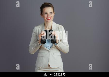 Glückliche junge kleine Geschäftsinhaberin in einem leichten Geschäftsanzug mit Fernglas vor grauem Hintergrund. Stockfoto