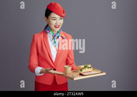 Lächelnde stilvolle Luft-Hostess asiatische Frau in rotem Rock, Jacke und Hut Uniform mit einem Tablett mit Essen isoliert auf grauem Hintergrund. Stockfoto