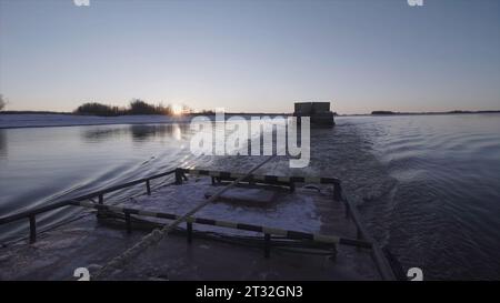 Blick auf das schwimmende Schiff mit Kabel auf dem Fluss. Clip. Schwimmende Binnenschiff hinter Schiff in Küstennähe mit Schnee an sonnigen Wintertagen. Lastkahn auf Kabel schwimmt dahinter Stockfoto